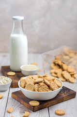 Small round crackers with oatmeal and sesame seeds in a white ceramic bowl on a wooden board on the table. in the background is a bottle of milk and a package of crackers.
