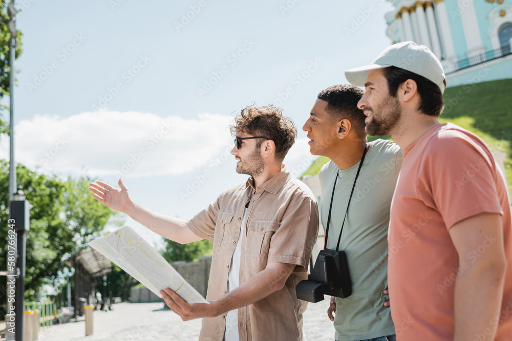 Wall mural side view of young guide with city map pointing with hand near multicultural travelers on andrews de
