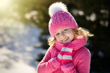Portrait of a pretty smiling blonde girl with blue eyes, dressed in a knitted hat with a pompom, sweater, scarf and pink mittens. The girl with golden hair folded her ankles and smiles