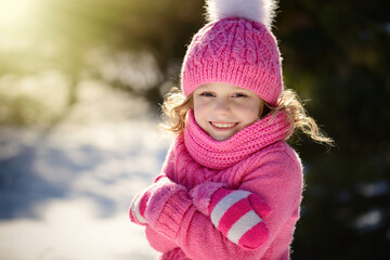 Portrait of a pretty smiling blonde girl with blue eyes, dressed in a knitted hat with a pompom,sweater, scarf and pink mittens. The girl with golden hair folded her arms hugging herself from the cold