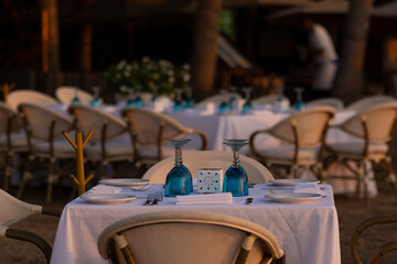 Served table with glasses, plates and white table cloth at the outdoor beach patio. Selective focus. Eating out, romantic dinner concept.