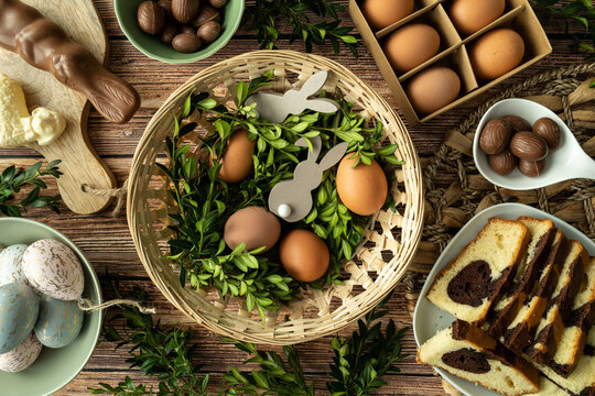 Traditional Easter Paschal wicker basket. Flat lay composition with eggs, boxwood branches, bunny decorations, chocolate candies and loaf cake.