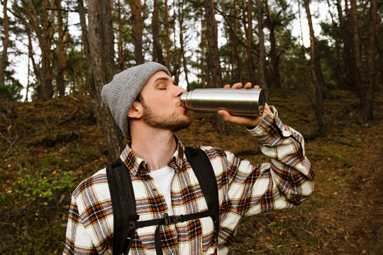 Young man drinking tea from thermos during hiking in green forest