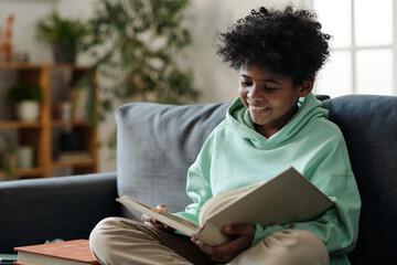 Smiling African American schoolboy in casualwear reading book while sitting on couch in living room and preparing for lesson of literature