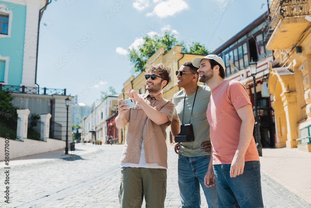 Wall mural smiling guide with mobile phone pointing with finger while leading tour for interracial men on podil