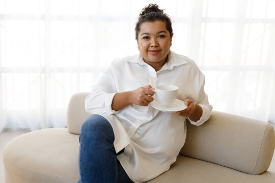 Cheerful young woman plus size sitting on couch, drinking coffee