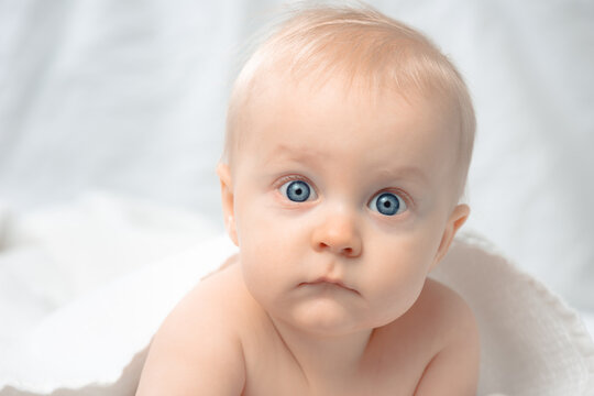 Cute baby with blue eyes - closeup portrait. Little boy looking at camera. Child lying on bed covered by bath towel.