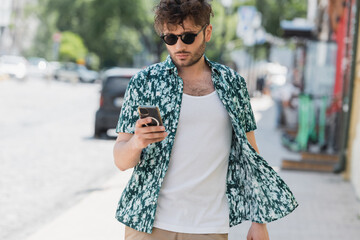 Brunette man in sunglasses using smartphone while walking on urban street.