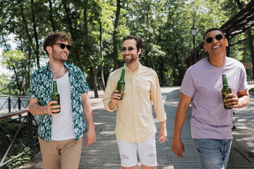 cheerful and trendy multiethnic friends holding fresh beer during walk in city park on summer day.