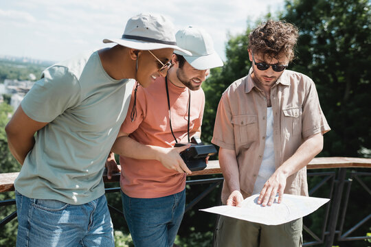 young tour guide in sunglasses showing map to interracial tourists in sun hats during excursion in city park.