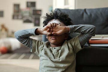 Offended or tired African American boy in grey pajamas hiding his face with hands while sitting by...