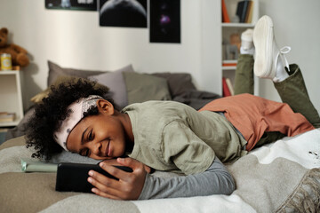 Cute smiling schoolboy relaxing on bed and watching online video on smartphone screen while enjoying staying at home on weekend