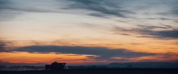 Silhouette of harvester machine to harvest wheat on sunset. Combine harvester driving on field on sunrise. Beautiful dawn sky above wheat field. Combine working in dusk. Wonderful twilight landscape. © Daniil