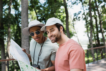 positive multiethnic tourists in sun hats looking at travel map in blurred park.