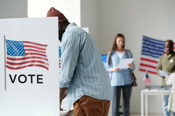 Side view of young African American male voter filling in ballot while standing by one of several...
