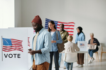 Group of young multicultural voters in casualwear standing in queue along vote booths in polling place and putting their ballots into boxes - obrazy, fototapety, plakaty