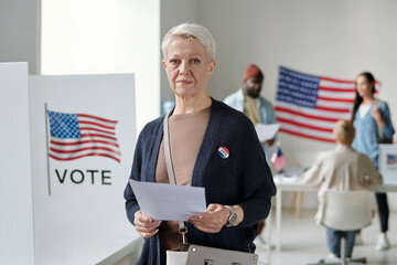 Confident mature blond female voter with ballot paper looking at camera while standing in polling...