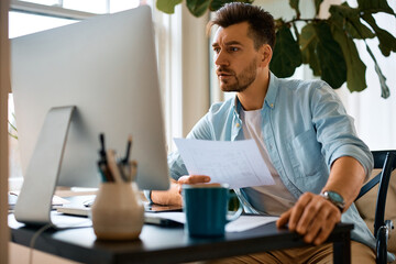 Mid adult businessman using computer while going through documents and working at home office.