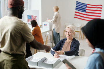 Happy blond mature woman passing ballot paper to young African American man standing by desk of...