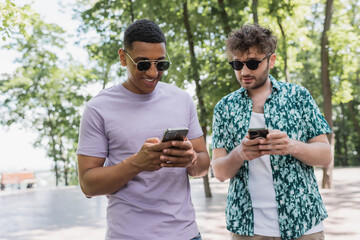 Smiling interracial friends in sunglasses using smartphones in park.