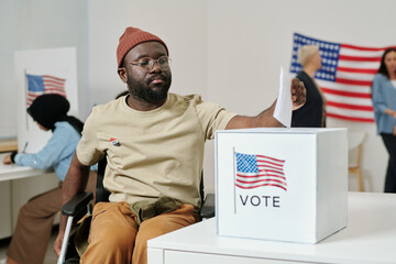 African American young man with disability sitting in wheelchair and putting ballot paper into box...
