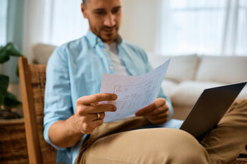 Close up of man reads document while using laptop at home.