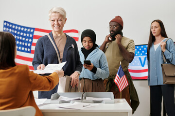 Queue of intercultural voters standing by desk of electoral commission while one of them taking...