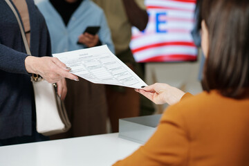 Close-up of young woman from electoral commission passing ballot paper to one of voters standing in...