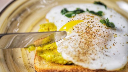 Cutting poached egg with runny egg yolk over bread toast with mashed avocado spread
