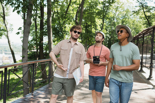 multicultural travelers in sun hats looking away during summer walk with tour guide in urban park.