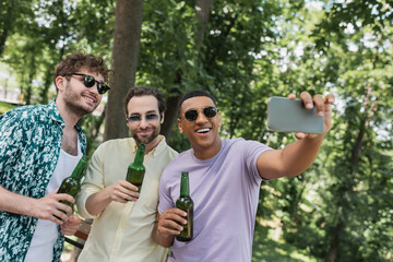 african american man holding beer and taking selfie with carefree friends in summer park.