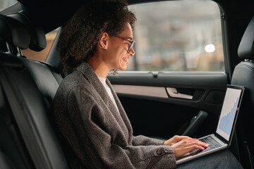 Smiling businesswoman manager working on laptop sitting car leather backseat on the way to office
