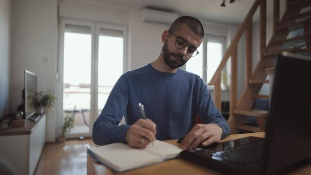 Professional focused and happy man working from home. Employee sit at desk at home working with laptop and papers, writes down ideas make notes and using phone