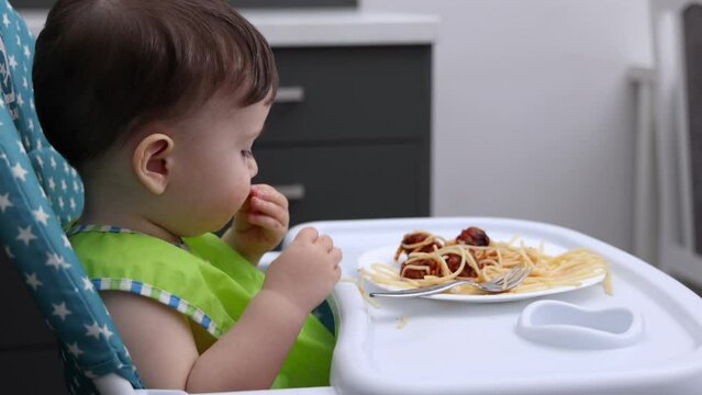 child baby boy eating pasta spaghetti with meatballs and tomatoes sauce sitting in high chair kitchen background.kid toddler putting food with both hand in mouth hungry cute infant.diversification
