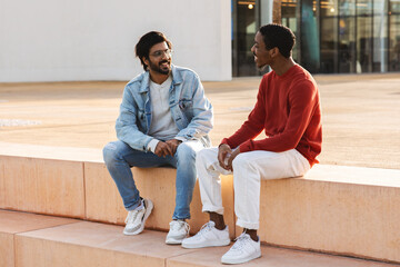 Happy young middle eastern and african american guys students in casual sit, have break and talk in...