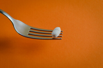 fork holding up a medicine pill on an orange background - concept for eating problems, medicine abuse, chemical feeding, health problems
