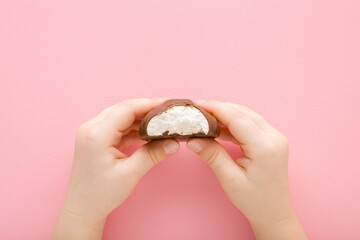 Little girl hands holding bitten white vanilla soft zephyr covered with dark brown chocolate glaze on light pink table background. Pastel color. Closeup. Children sweet snack. Top down view.