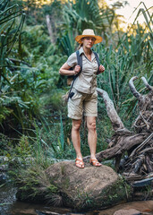 Young woman in beige shirt, shorts and straw haw, backpack on shoulders, hiking rainforest jungle, small creek near