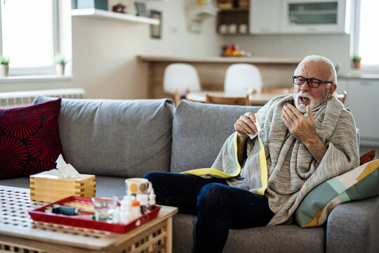 A Senior Caucasian Man Is Lying In Bed With A Bad Cold And Is Blowing His Nose Using A Napkin. An Older Ill Man With Temperature Is Lying In His Bed Covered With A Blanket.