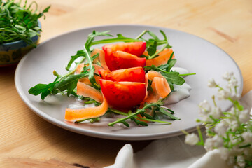 Rocket salad with tomatoes, carrots and yogurt sauce on a grey plate, with flowers and kitchen towel over a wooden table