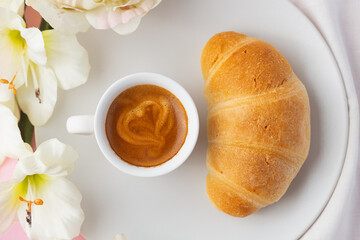 Top close upview of croissant on a white plate with a cup of espresso coffee with a heart on it, flowers and pink background