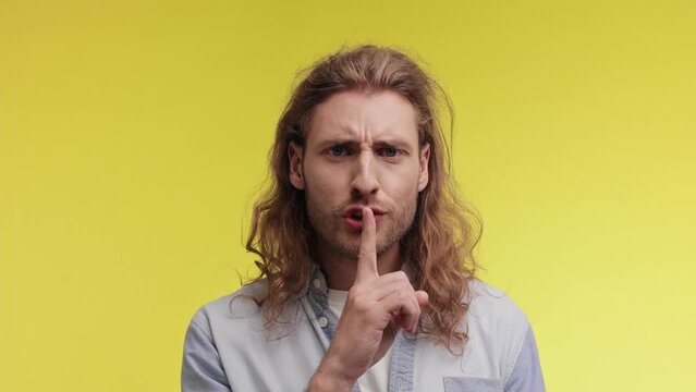 Portrait of Secret young man wears denim shirt look aside say hush be quiet with finger on lips shhh gesture close his mouth isolated on yellow color background studio. People emotion concept.