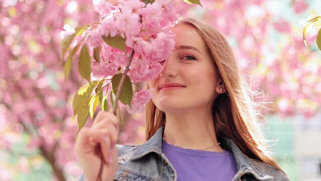 Close up portrait young face woman with beautiful eyes looking and smiling at camera on blurred Sacura tree background in the city street. Happy Caucasian girl with Cherry blossom trees. Slow motion.