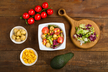 Vegetable salad with champignon corn and cherry tomato with avocado in a white plate on a wooden table next to all the ingredients.