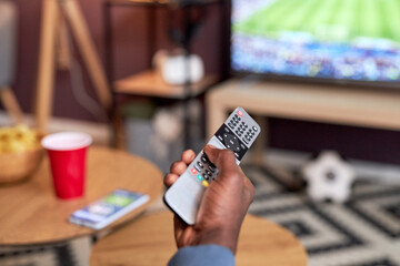 Close up of black young man watching football match at home with focus on male hand holding TV remote, copy space