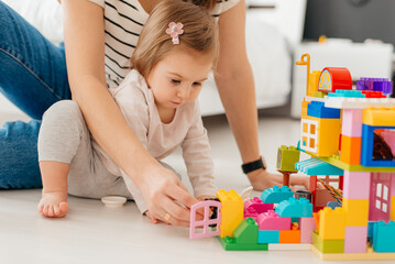 Laughing mother and little daughter playing colorful blocks, constructing tower, sitting on warm floor with underfloor heating, family enjoying leisure time together