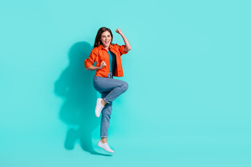 Full length photo of sweet lucky lady wear orange shirt jumping high rising fists empty space isolated teal color background