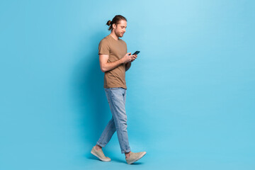 Full length photo of sweet cute man wear beige t-shirt walking empty space chatting modern gadget isolated blue color background