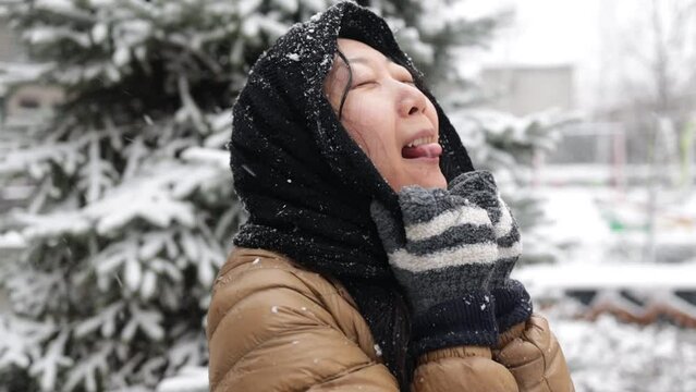 Funny asian girl is catching snowflakes by mouth at winter, portrait of charming girl outdoors