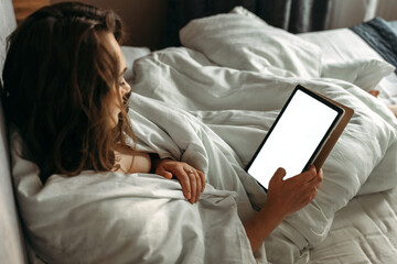A young woman lies in bed in the morning, looks at the tablet.